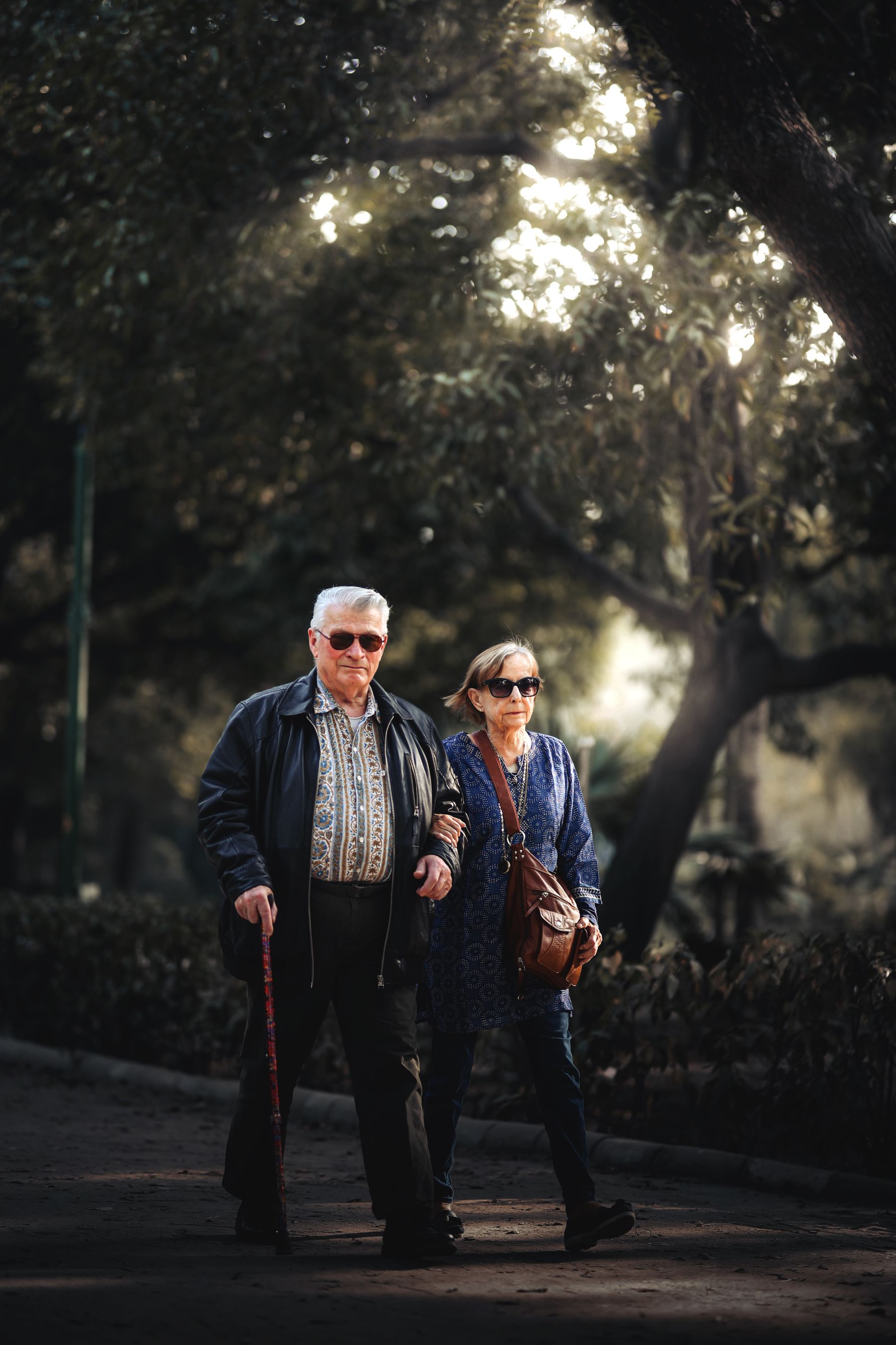 Illustrative photograph: An older couple in smart casual clothes and sunglasses walking together through a leafy city park.