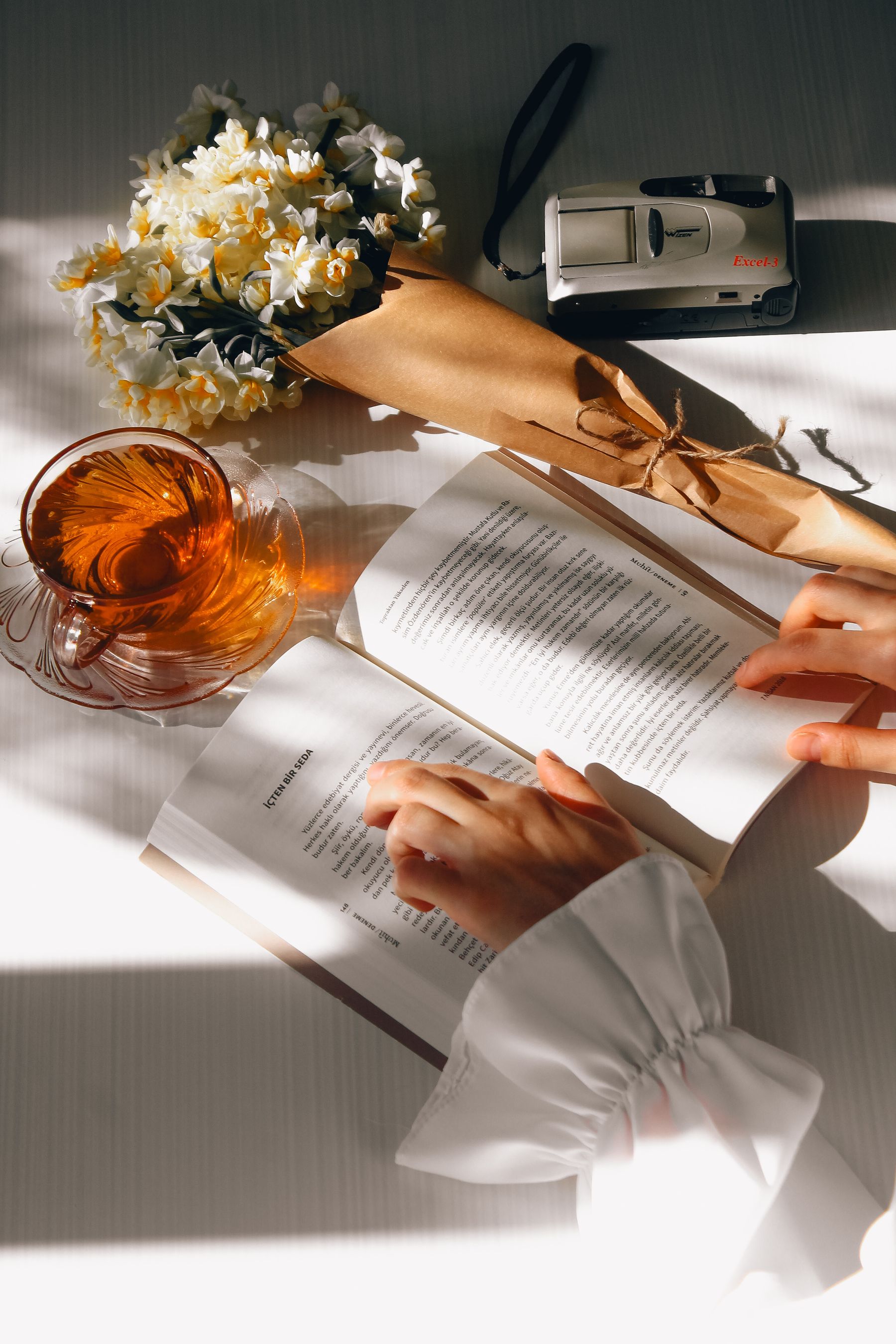 Illustrative photograph: An open book beside tea and flowers in natural window light.