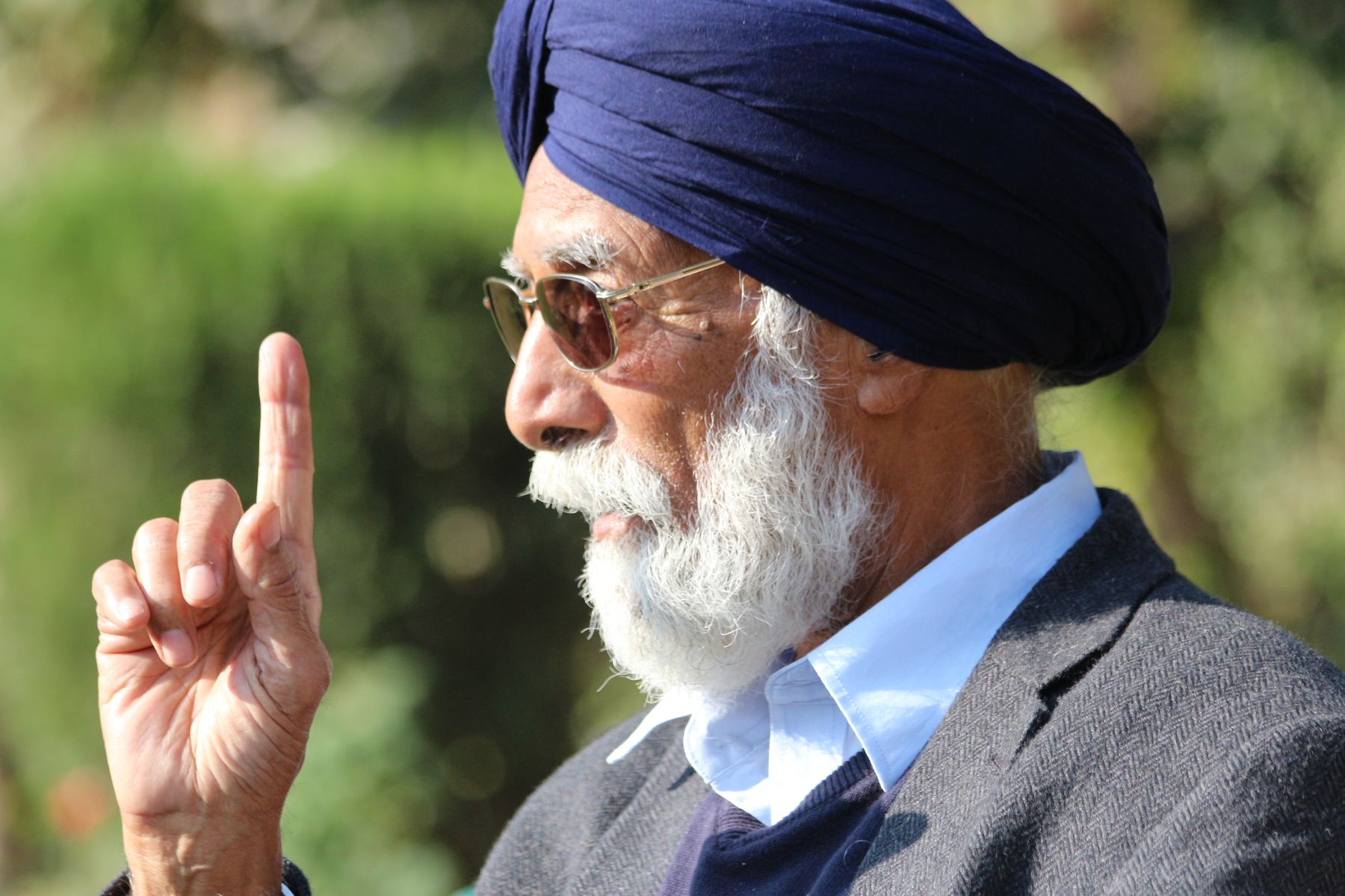 Illustrative photograph: An older man in a navy turban and tailored blazer talking outdoors against soft green trees.