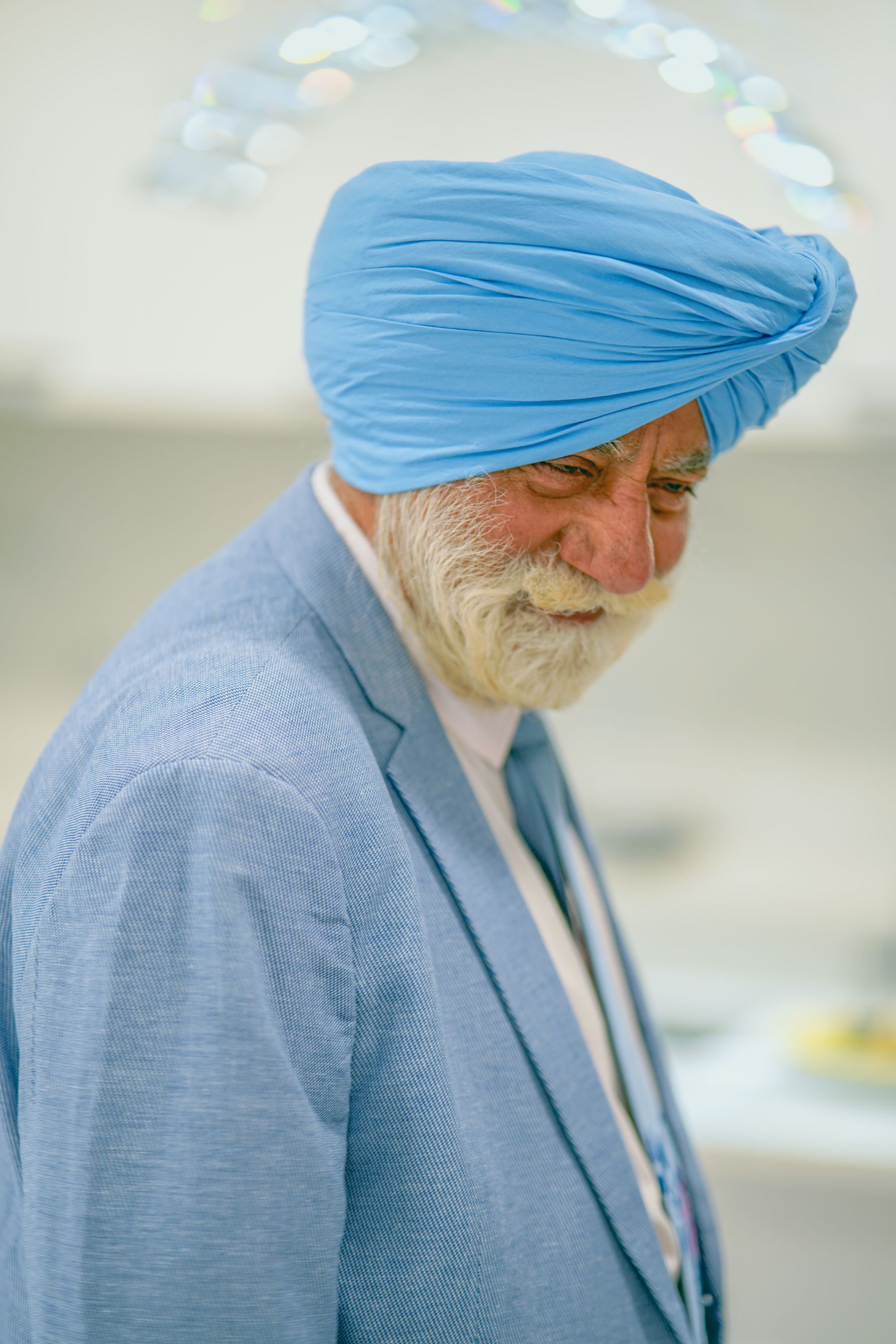 Illustrative photograph: An older Sikh man in a blue turban and smart blue suit sharing a warm smile indoors.